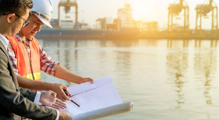 Strategic Port Planning at Sunrise Two professionals, including an engineer in a safety vest and helmet, analyzing construction plans against the backdrop of a port with cranes and water in the golden morning sunlight.
