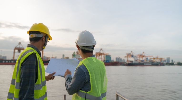 On-Site Planning with a Port View Two engineers wearing safety vests and helmets stand at a harbor reviewing construction plans, with cranes and container ships in the background.