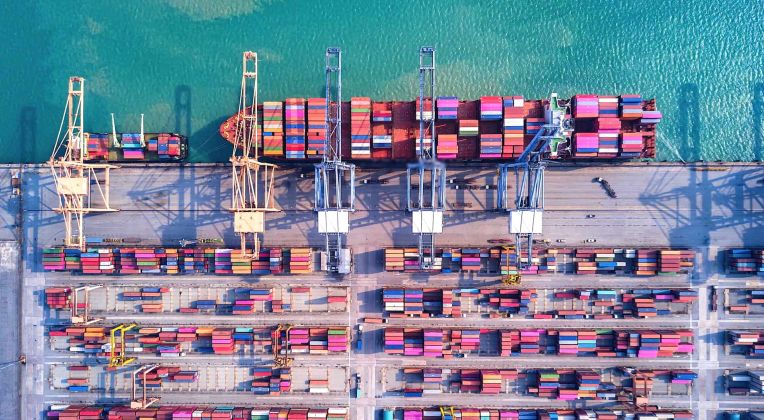 Port Operations from a Bird's Eye View Aerial view of a container port with stacked containers, large cranes, and a cargo ship in turquoise water.